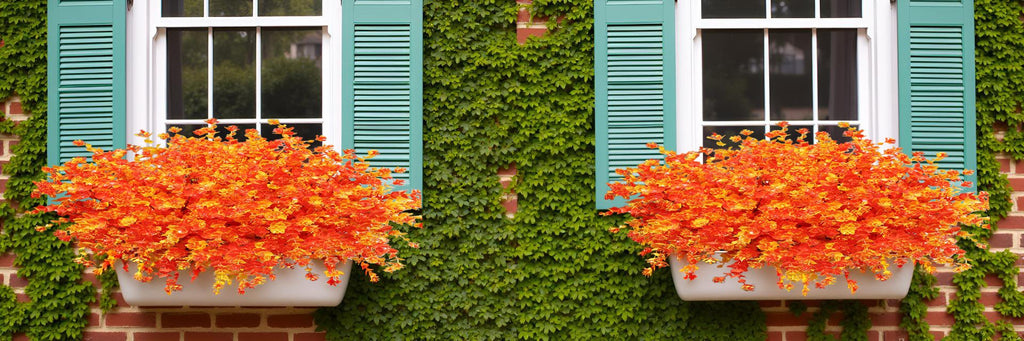 Two white window boxes filled with vibrant orange and yellow artificial fall bushes, set against a brick wall with green ivy and teal shutters.