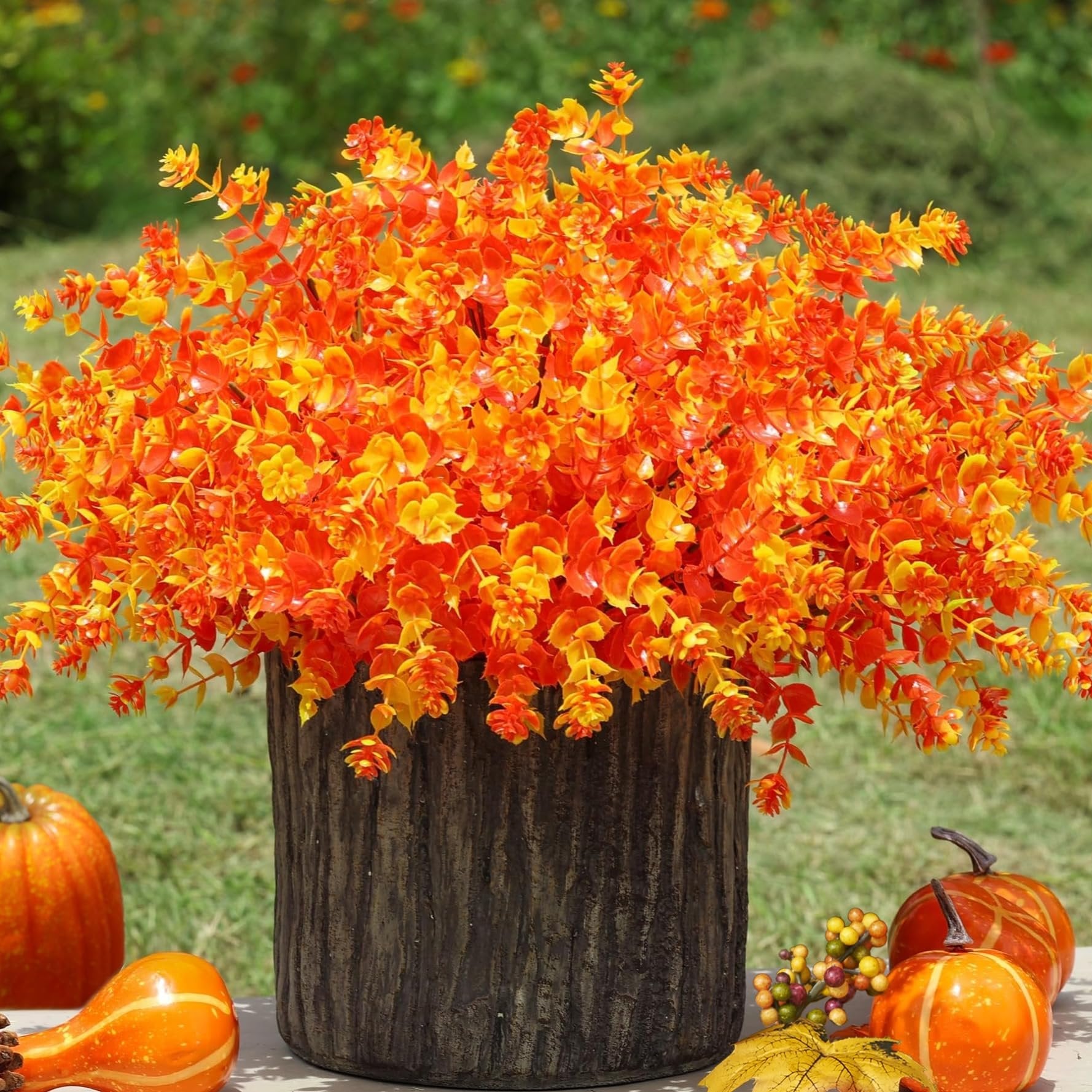 Artificial fall eucalyptus bushes in orange and yellow hues in a dark wood-textured pot, surrounded by pumpkins and gourds.
