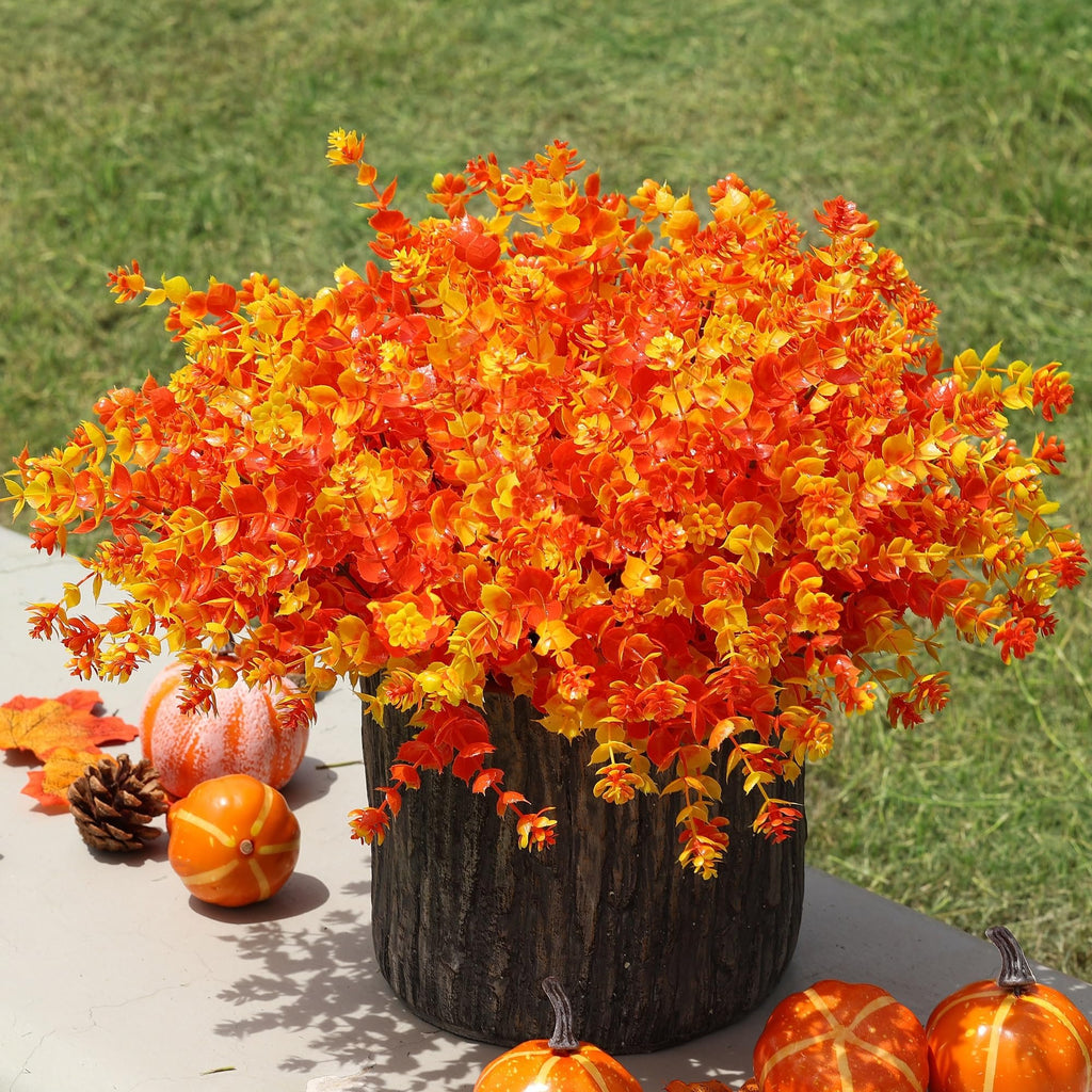 Artificial fall eucalyptus bushes in a wood-textured pot with decorative pumpkins and a pinecone on a ledge.