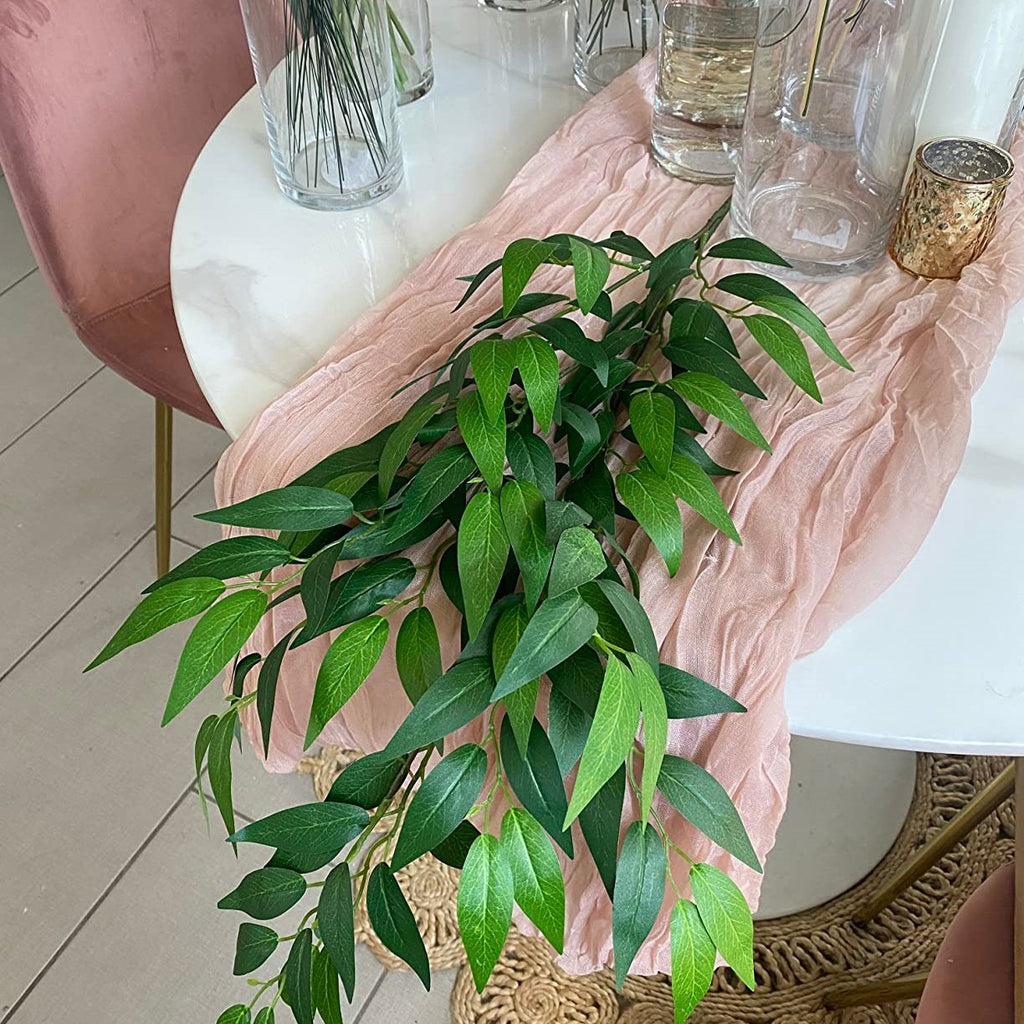 Artificial Italian ruscus greenery stem with numerous green leaves draped over a pink runner on a white table.