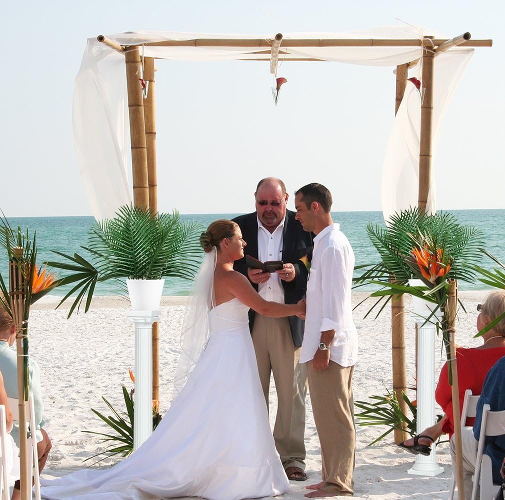 Beach wedding ceremony with a bride and groom exchanging vows under a bamboo arch, decorated with artificial palm leaves and tropical flowers.