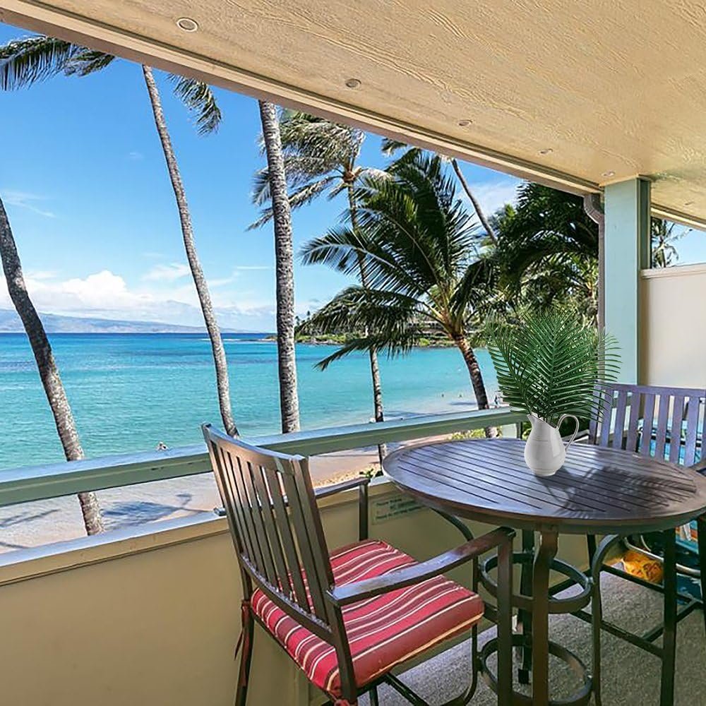 Balcony view of a tropical beach with palm trees and turquoise water, featuring a table with a vase of artificial palm leaves.