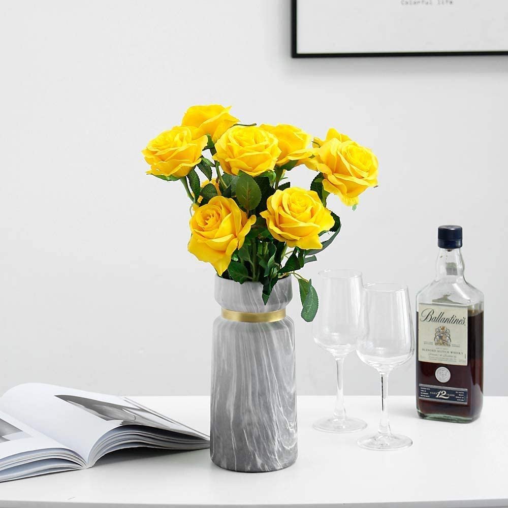A vase of 12 yellow silk roses with green leaves, placed on a white table next to an open book, two wine glasses, and a bottle of Ballantine's Scotch Whisky.