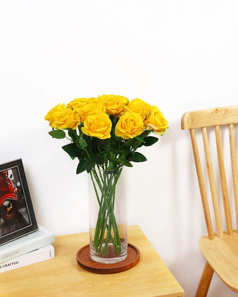 A dozen yellow silk roses in a clear glass vase on a wooden table next to a chair.
