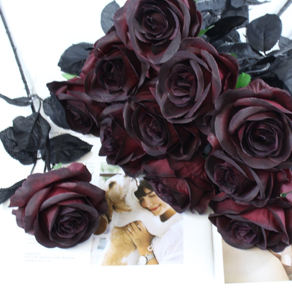 Burgundy artificial roses with black leaves arranged on a white surface with a magazine spread showing a woman and dog