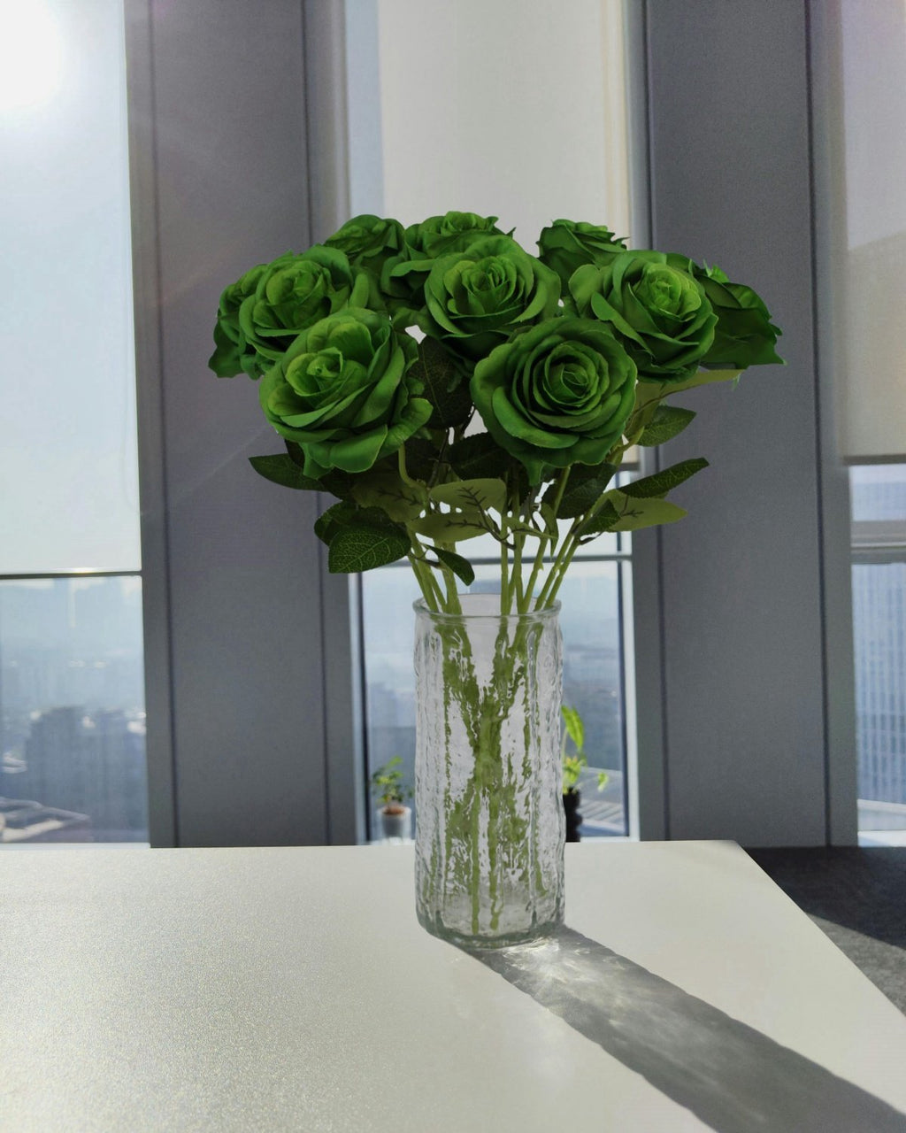 Bouquet of 12 green artificial roses in a textured glass vase on a white table, with a city view through windows in the background.