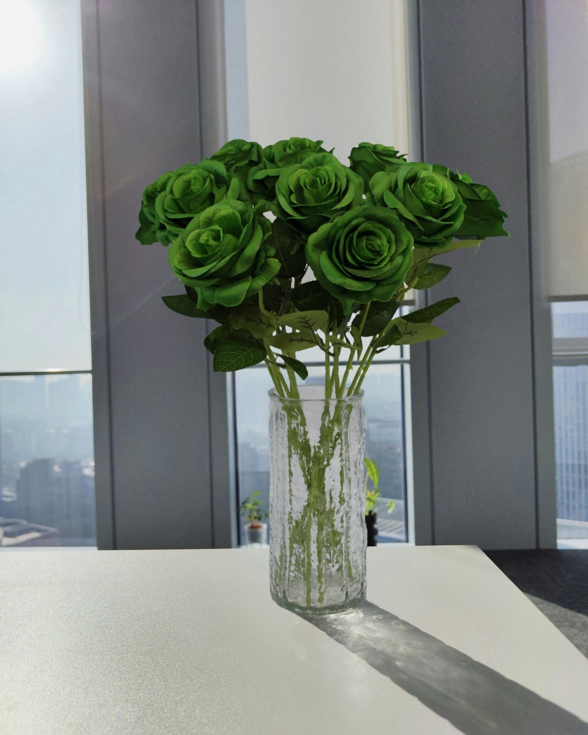 Bouquet of 12 green artificial roses in a textured glass vase on a white table, with a city view through windows in the background.