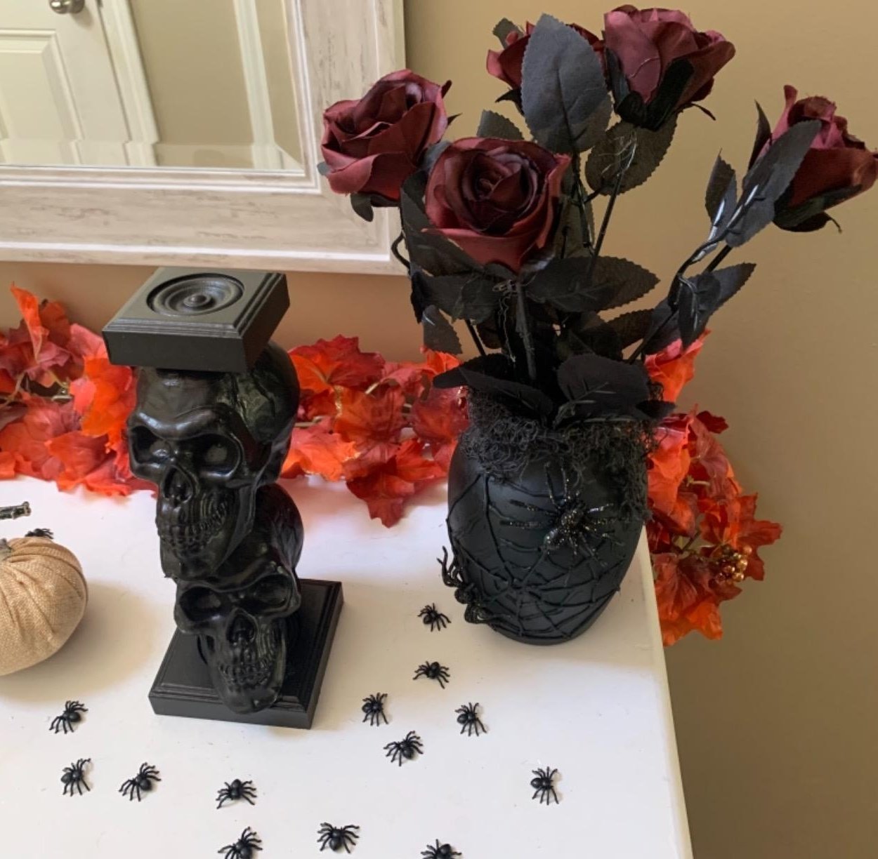 Halloween decor featuring a black skull candle holder, a vase with dark red artificial roses, and scattered plastic spiders on a white surface.
