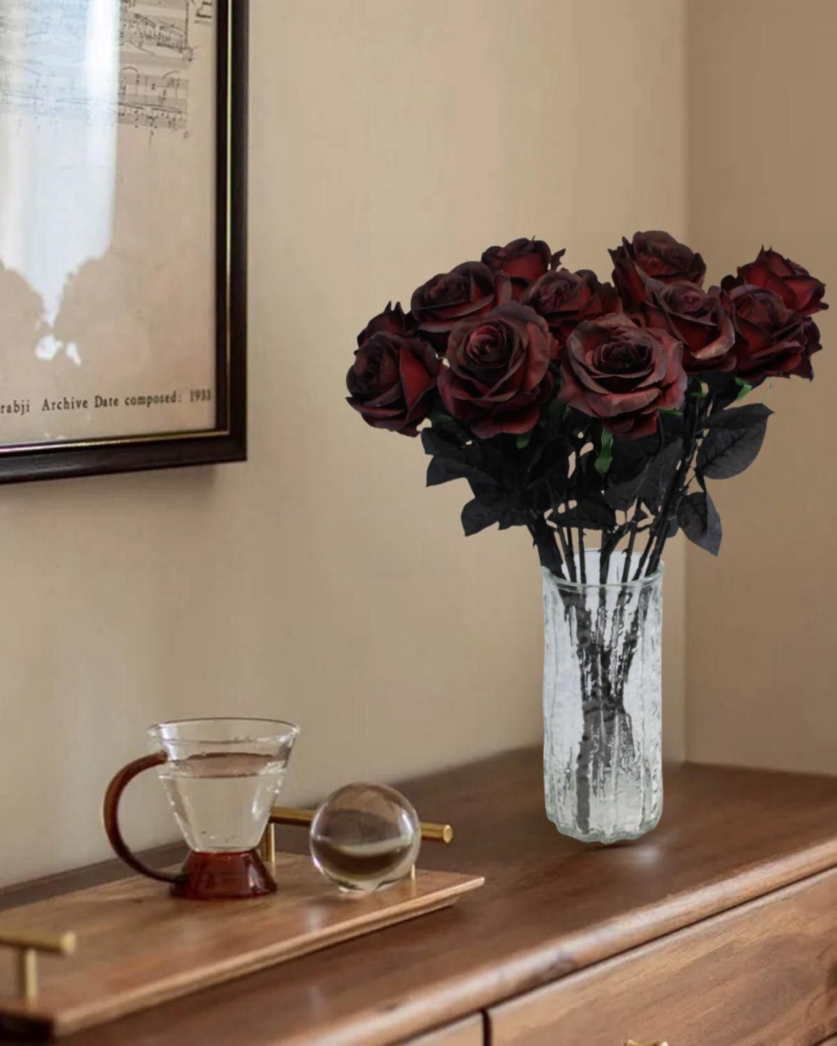 Bouquet of 12 burgundy artificial roses in a glass vase on a wooden dresser next to a glass of water.
