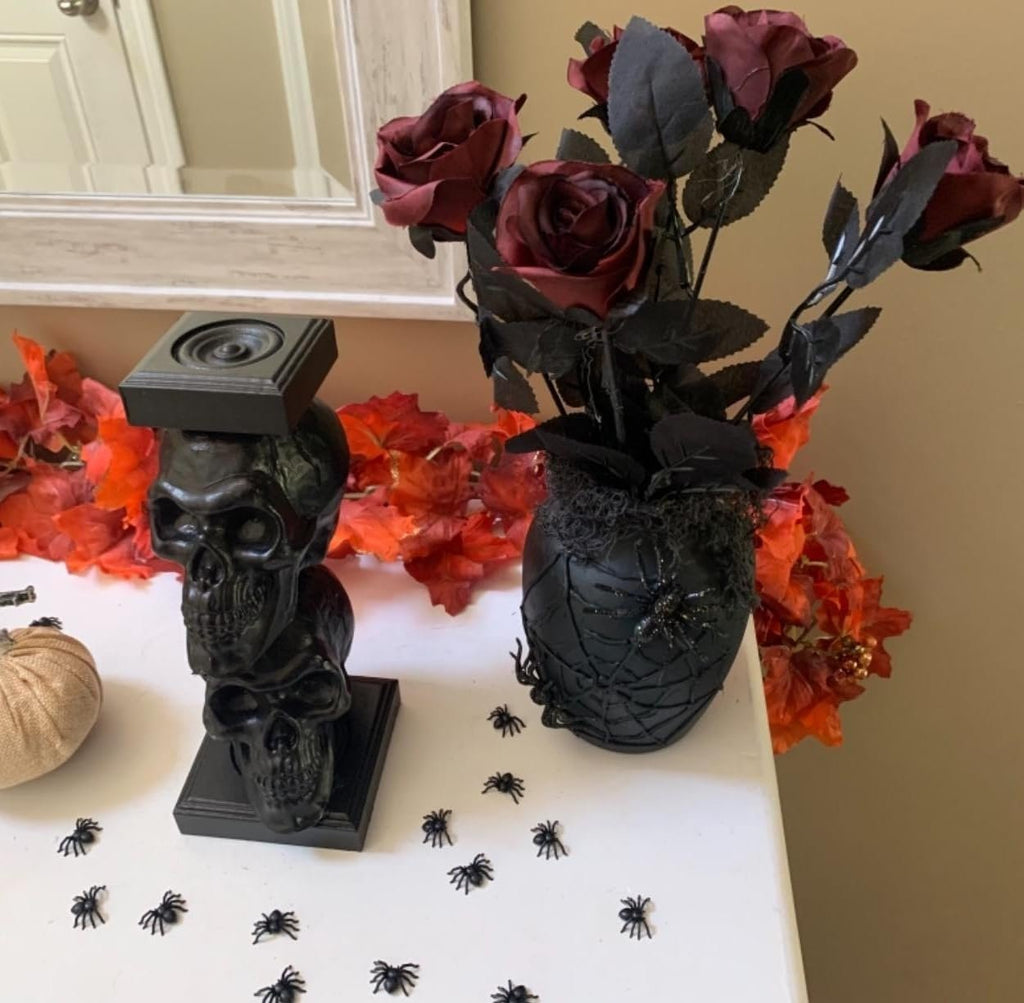Burgundy artificial roses in a black spiderweb vase with a stacked skull candle holder and small plastic spiders on a white surface.