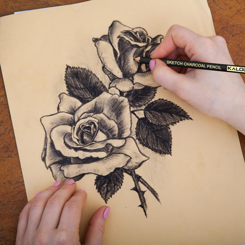 Hands drawing a charcoal sketch of roses on tan paper with a charcoal pencil.