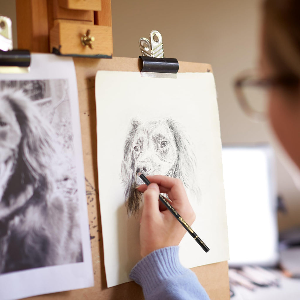 Artist's hand sketching a dog portrait with a charcoal pencil on paper, reference photo visible