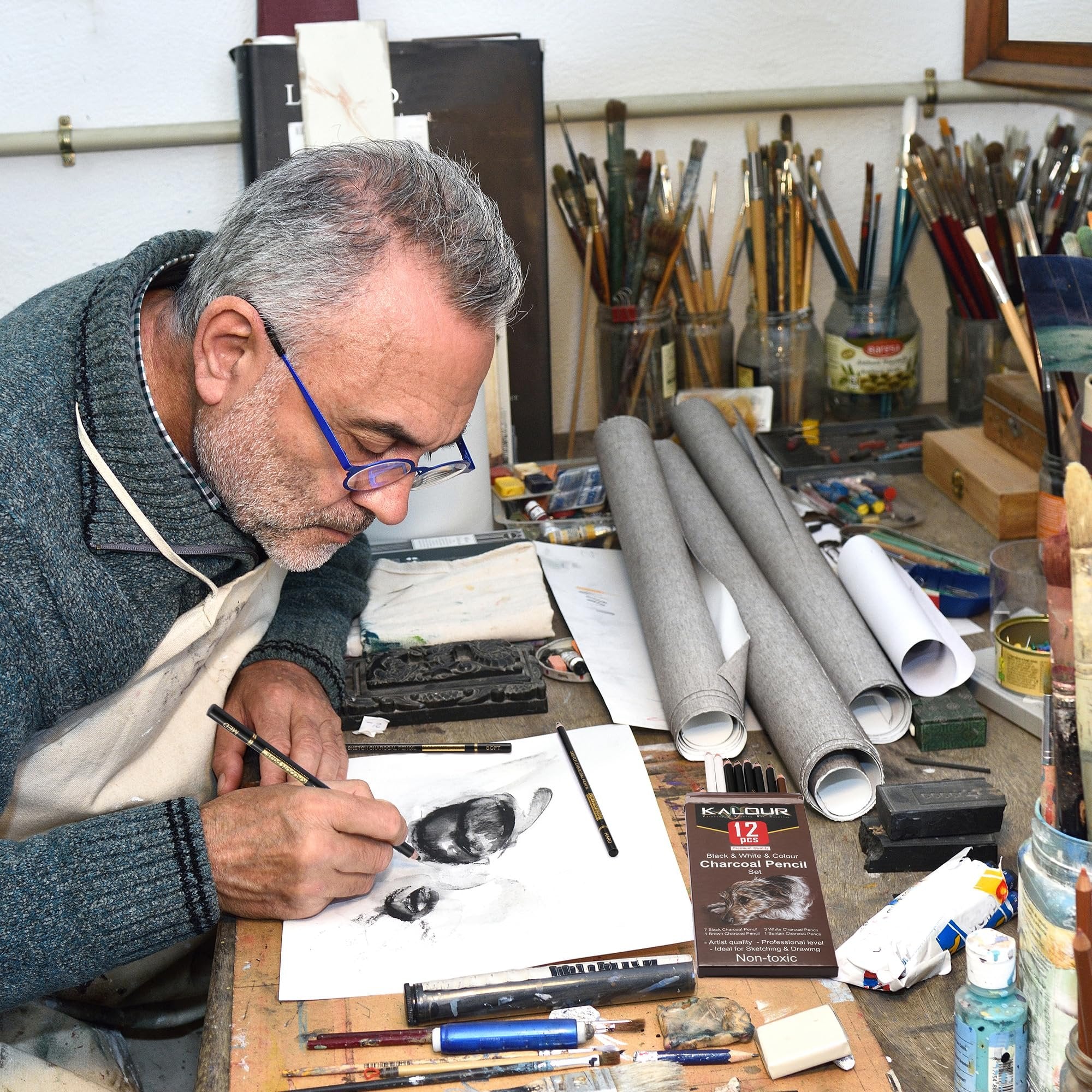 Artist sketching with charcoal pencils at a cluttered desk with art supplies and rolled canvases.
