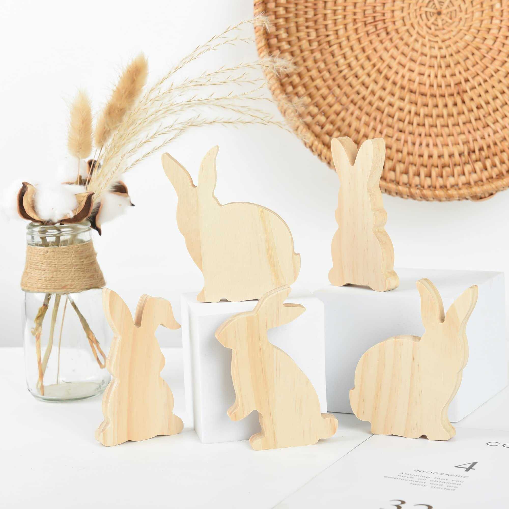 Five unfinished wooden bunny cutouts arranged on a white surface with decorative dried grasses and a woven basket in the background.