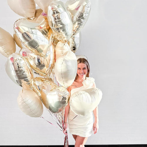 Woman holding a large bouquet of cream white, white, and light gold heart-shaped foil balloons against a white brick wall.