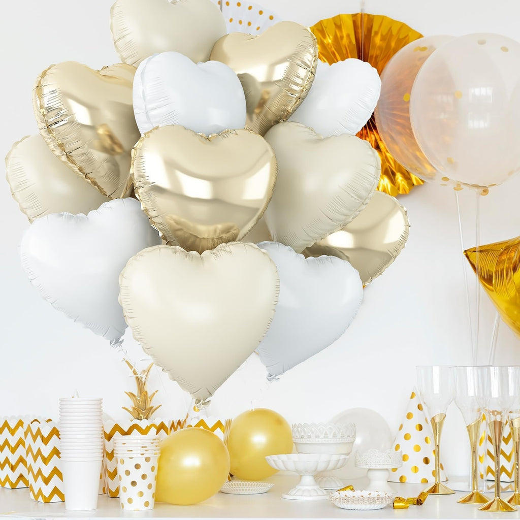A bouquet of cream white, white, and light gold heart-shaped foil balloons arranged on a white surface with party decorations.