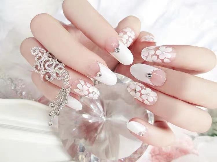 Close-up of a hand with white and clear floral-patterned artificial nails adorned with rhinestones, holding a decorative silver ring with tassels.