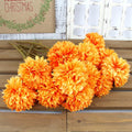 A bunch of 12 orange artificial chrysanthemum flowers with green stems lying on a wooden surface, with a Christmas-themed burlap sign in the background.