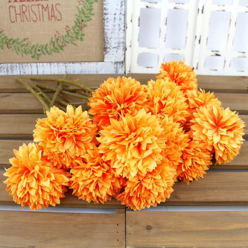 A bunch of 12 orange artificial chrysanthemum flowers with green stems lying on a wooden surface, with a Christmas-themed burlap sign in the background.