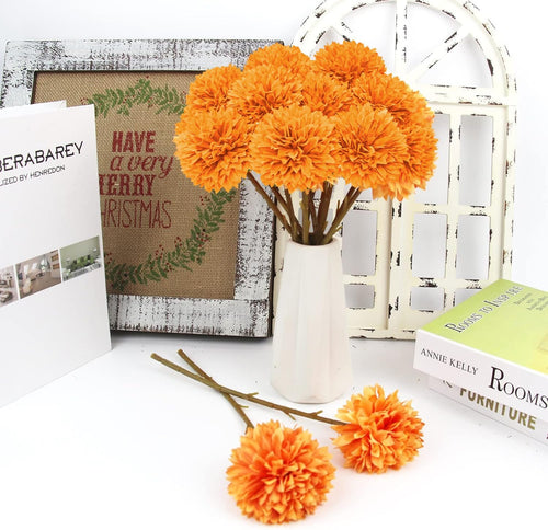 A bouquet of 12 orange artificial chrysanthemum flowers in a white vase, with two flowers and stems on the side, next to decorative items.