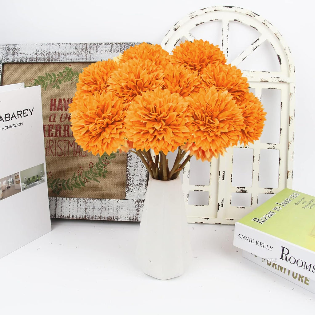 A bouquet of 12 orange artificial chrysanthemum flowers in a white vase, with decorative items in the background.