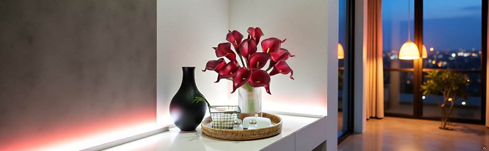 Bouquet of dark red calla lilies in a vase on a shelf with a black vase and decorative items, with a city view at dusk through a window.