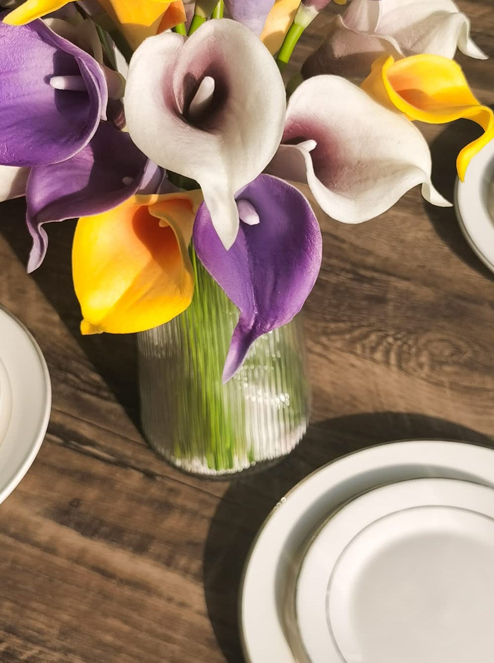 Close-up of a bouquet of colorful artificial calla lilies in a glass vase on a wooden table with plates.