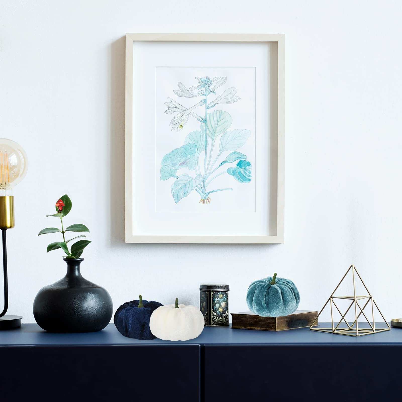 Velvet pumpkins in navy, white, and teal arranged on a dark blue cabinet with a framed botanical print above.