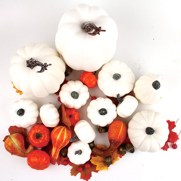 Assorted white and orange artificial pumpkins and gourds with fall leaves and acorns on a white background.