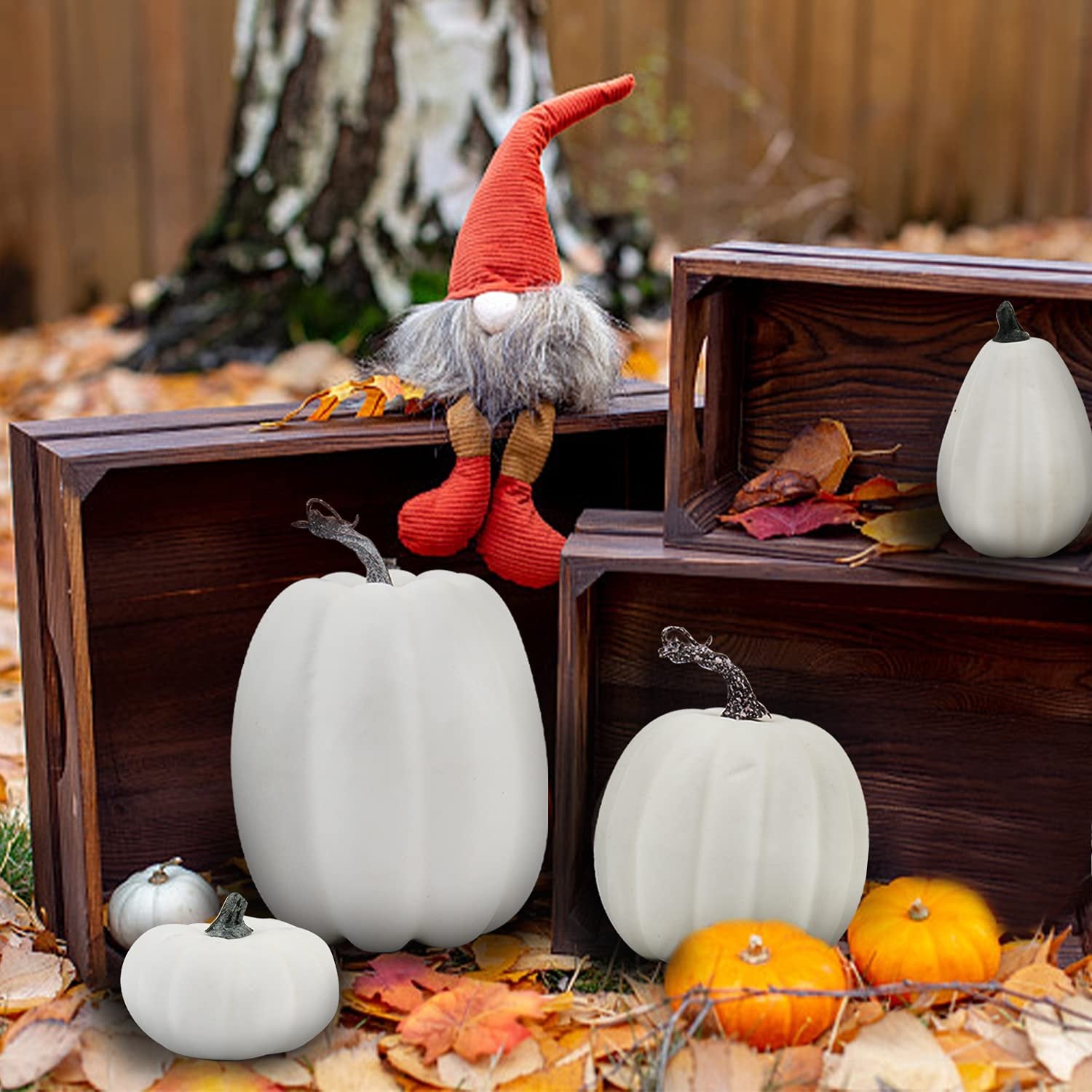Assorted white and orange artificial pumpkins arranged with wooden crates and a gnome figure on fallen leaves.