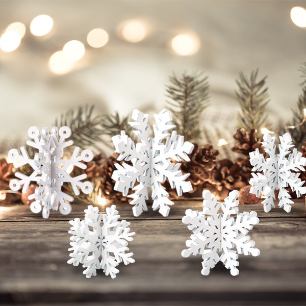 Five white wooden 3D snowflake decorations arranged on a rustic wooden surface with pine cones and blurred fairy lights in the background.