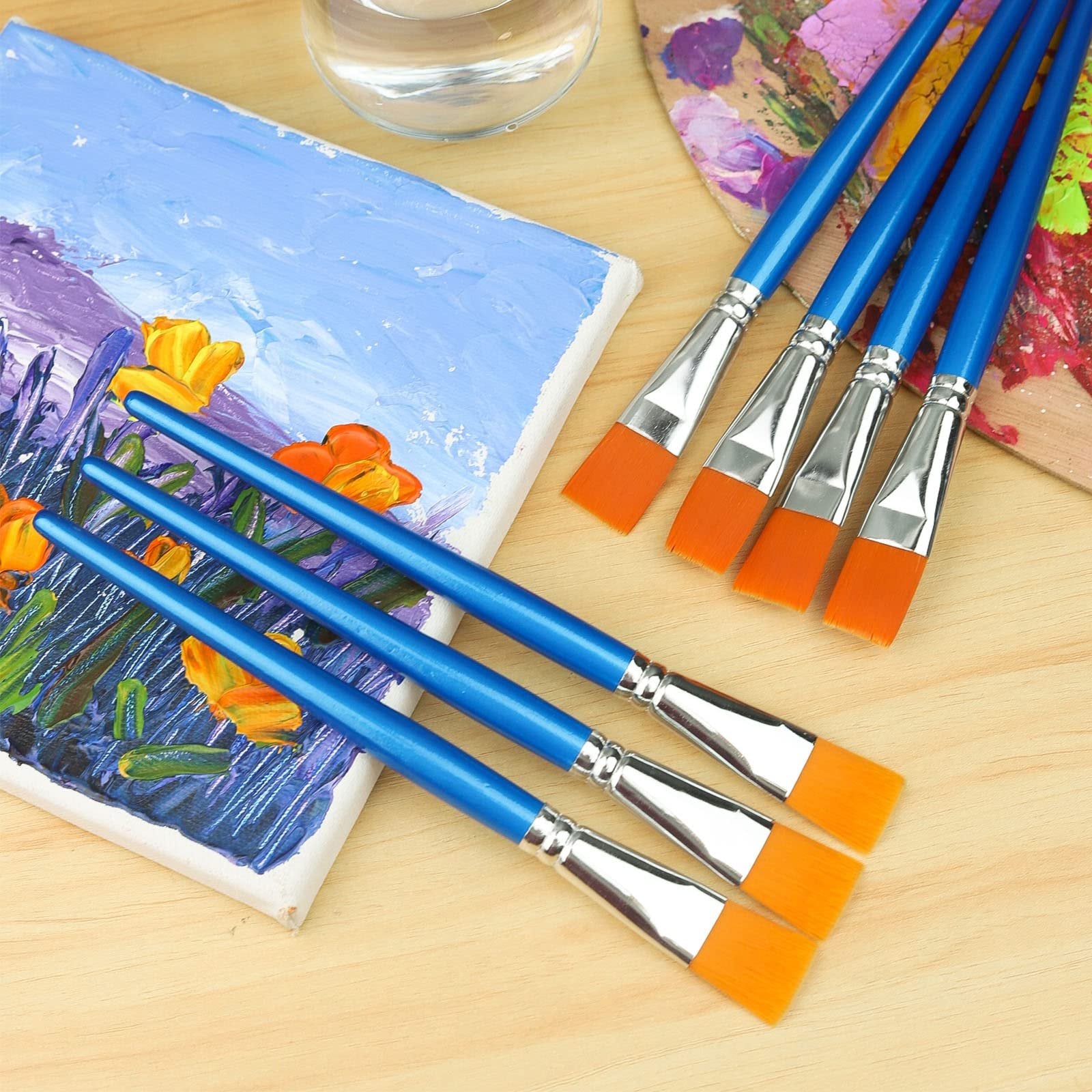 Flat paint brushes with blue handles and orange bristles arranged next to a small painting and a palette on a wooden surface.