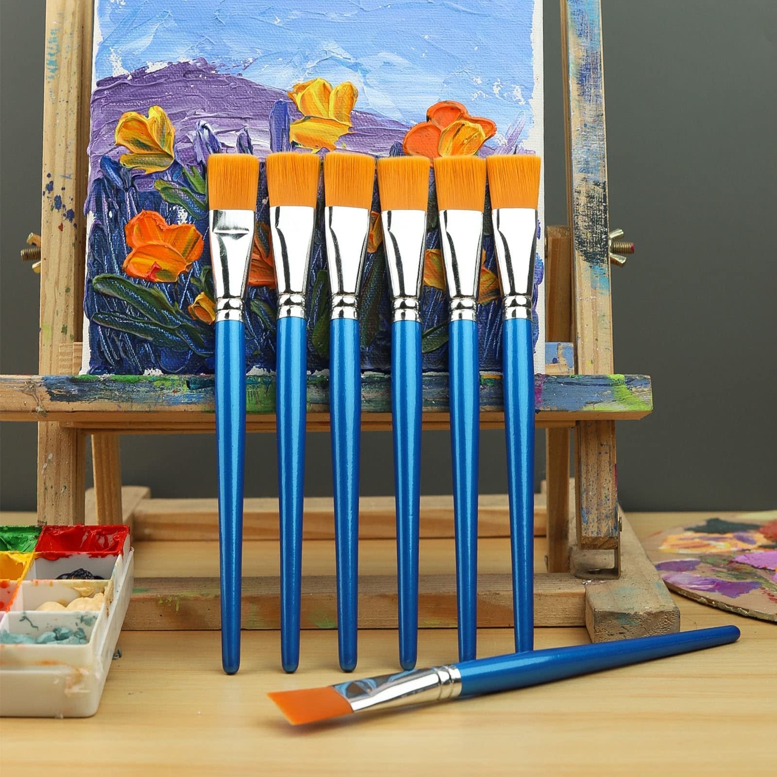 Six flat paint brushes with blue handles and orange bristles stand on a wooden surface in front of a small easel displaying a colorful painting. One brush lies in the foreground.