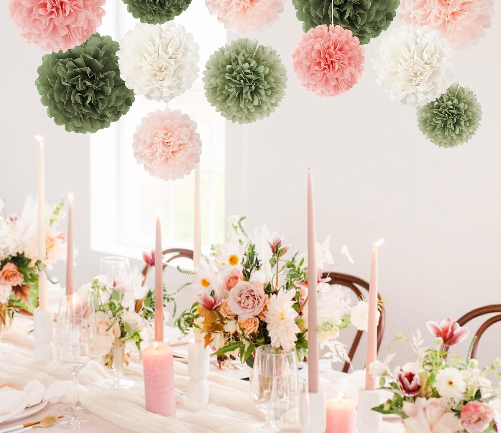 Pink, sage green, and white tissue paper pom poms hanging above a decorated table setting with flowers and candles.
