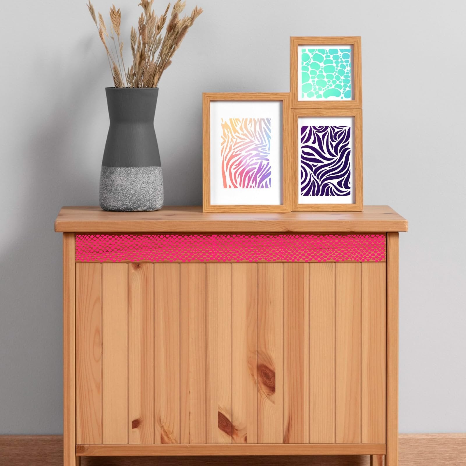 Three framed art prints with abstract patterns displayed on a wooden cabinet next to a vase of dried grass.