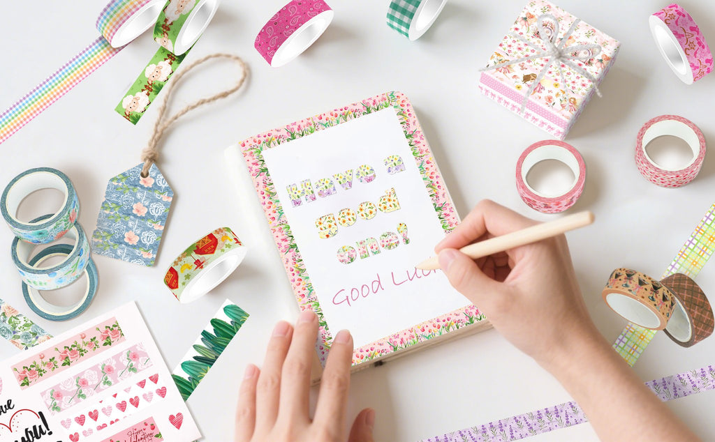 Hand writing 'Good Luck' on notebook surrounded by various floral and patterned washi tape rolls