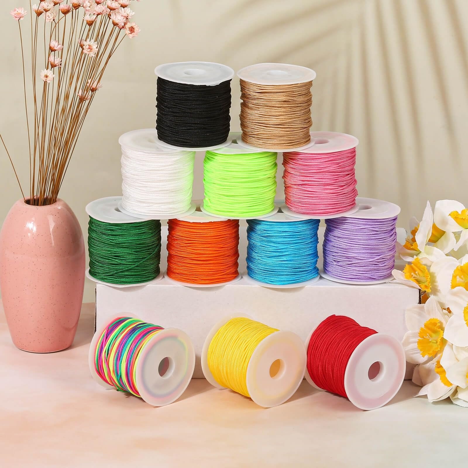 Assortment of 12 spools of nylon cord in various colors, including black, tan, white, neon green, pink, green, orange, blue, purple, multi-color, yellow, and red, arranged on a surface with decorative flowers.