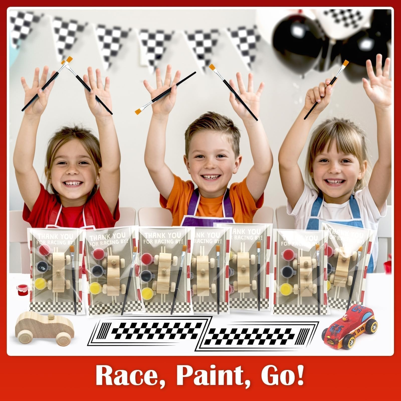 Three smiling children hold paintbrushes up, with unfinished wooden race car painting kits laid out in front of them.