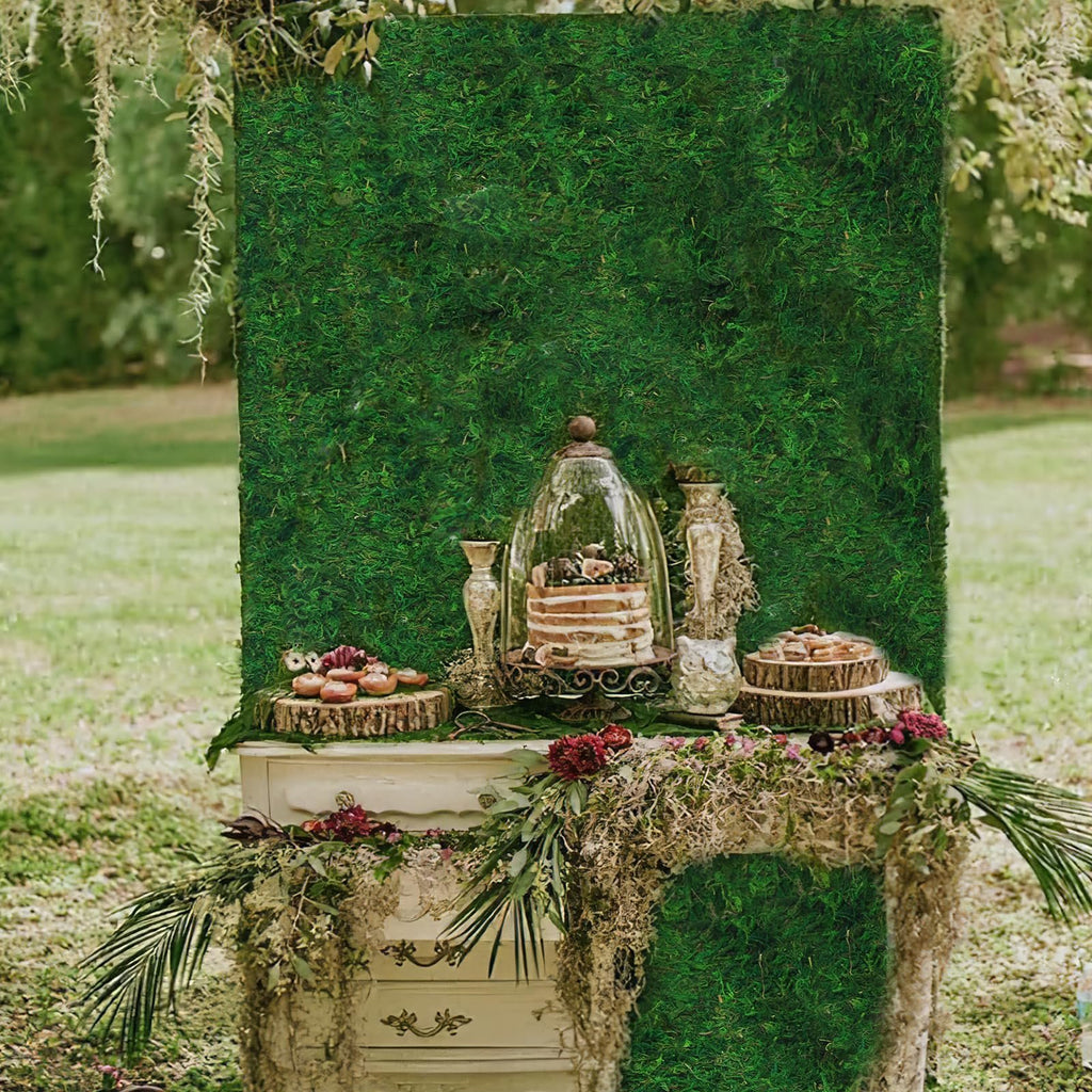 Wedding dessert table setup with a moss backdrop, cake under a cloche, and decorative elements.