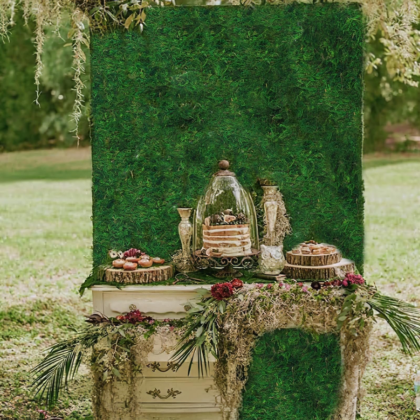 Wedding dessert table setup with a moss backdrop, cake under a cloche, and decorative elements.