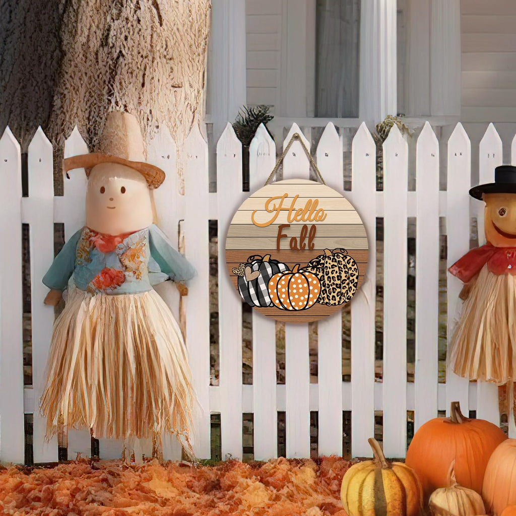 Round wooden fall door sign with pumpkins and 'Hello Fall' text, hanging on a white picket fence with scarecrows and pumpkins.