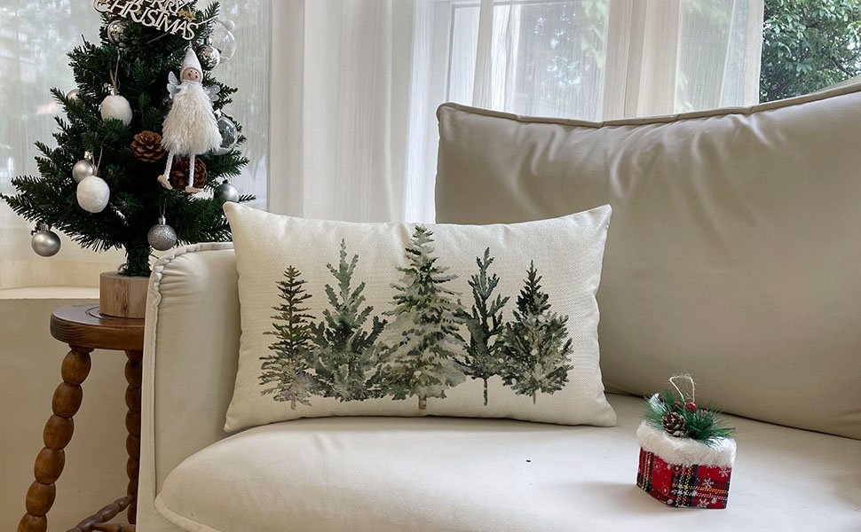 Rectangular throw pillow cover with a watercolor forest of pine trees, placed on a sofa next to a small decorated Christmas tree.