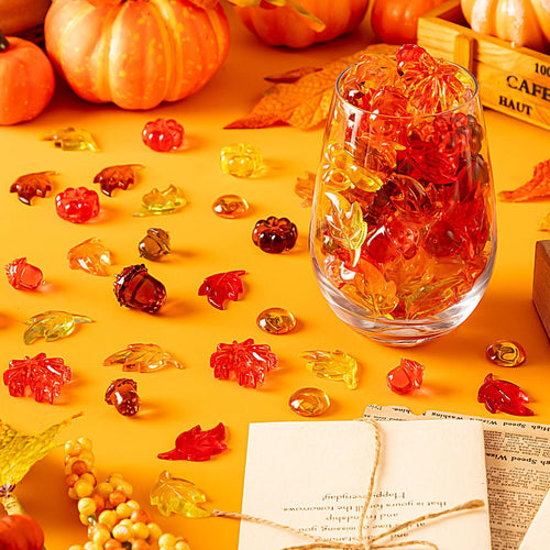 Acrylic fall vase filler set with pumpkins, leaves, acorns, and gems scattered on an orange surface, with a glass filled with the filler.