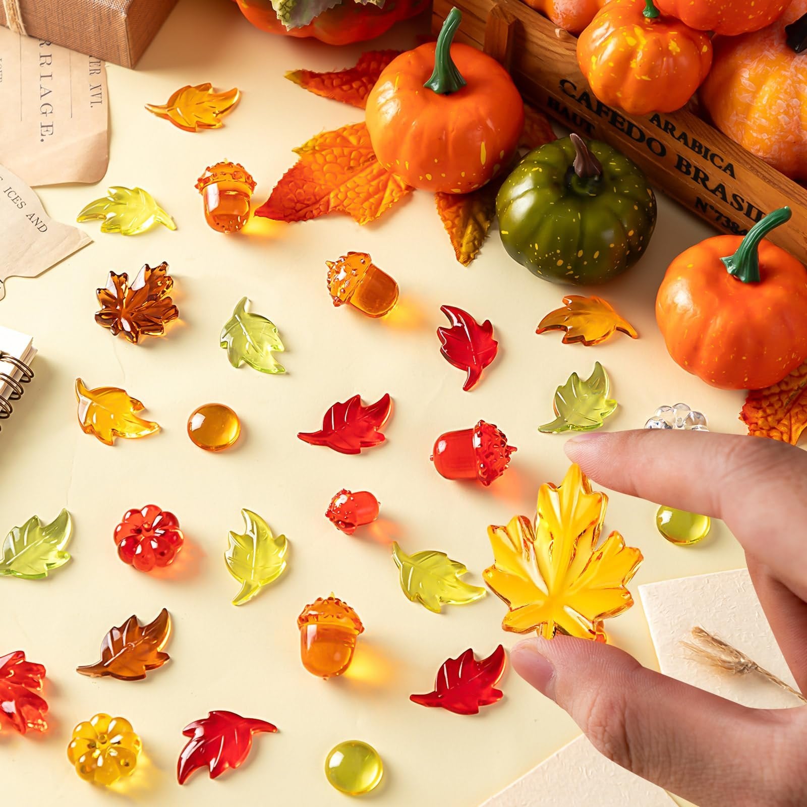 Acrylic fall vase filler pieces including leaves, acorns, pumpkins, and gems scattered on a surface with decorative pumpkins and a wooden crate.