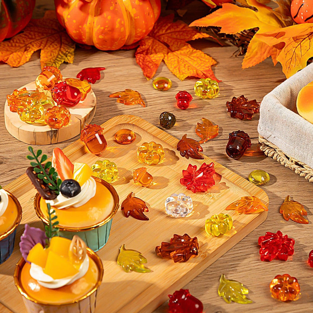 Assortment of acrylic fall-themed decorations including pumpkins, leaves, acorns, and gems scattered on a wooden surface and a bamboo tray, with cupcakes in the foreground.