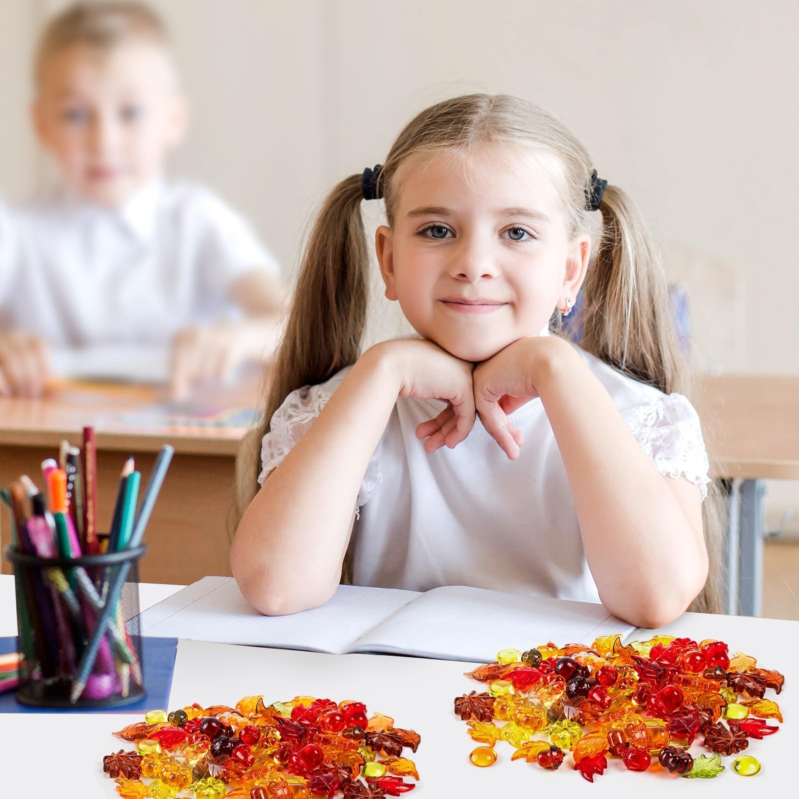 Young girl with pigtails resting chin on hands, looking at camera, with piles of colorful acrylic fall-themed vase filler on table in foreground.