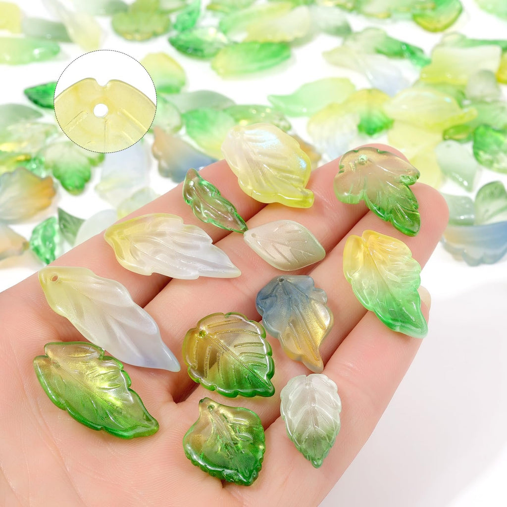 Hand holding assorted glass leaf beads in green, yellow, and white hues, with a close-up inset showing bead detail.