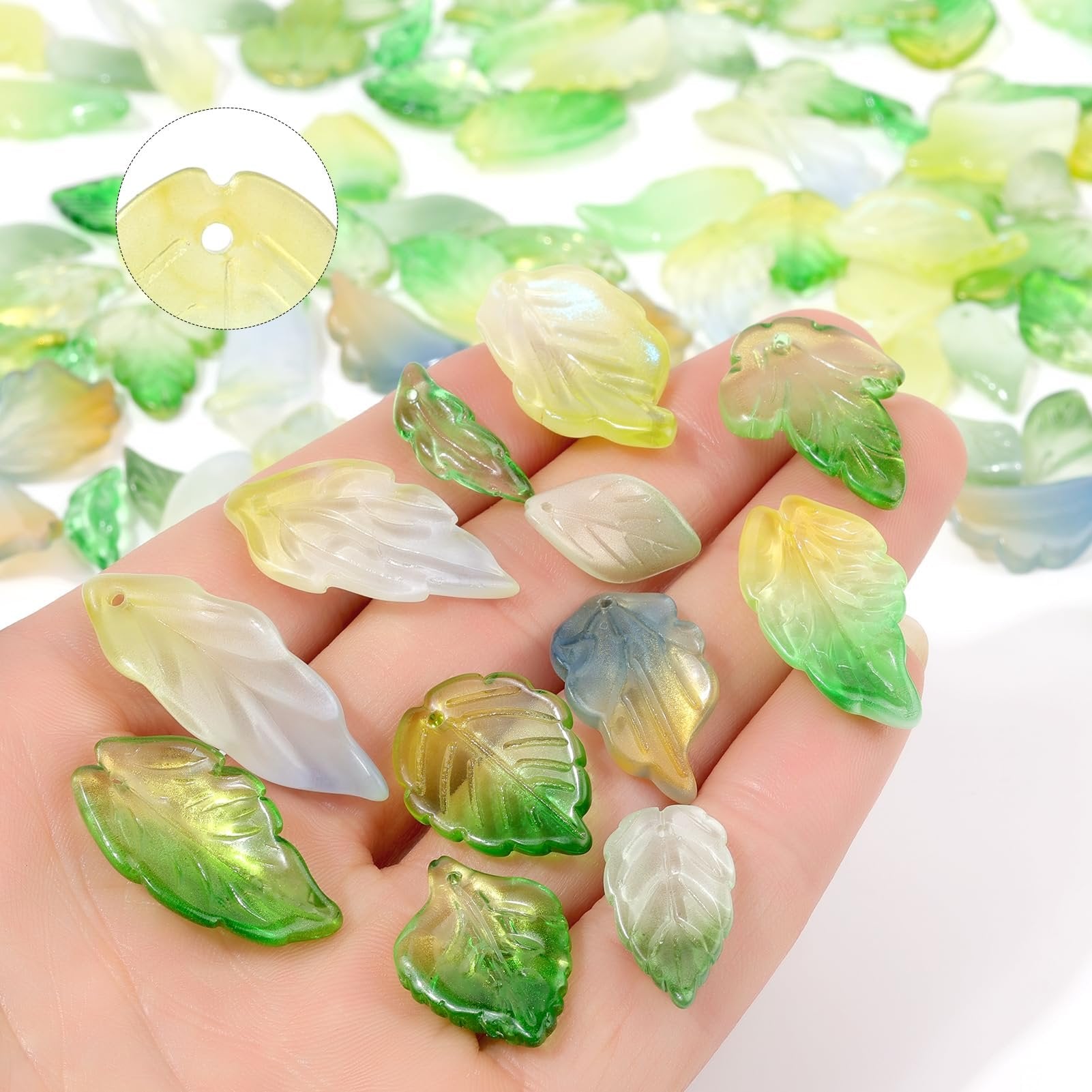 Hand holding assorted glass leaf beads in green, yellow, and white hues, with a close-up inset showing bead detail.