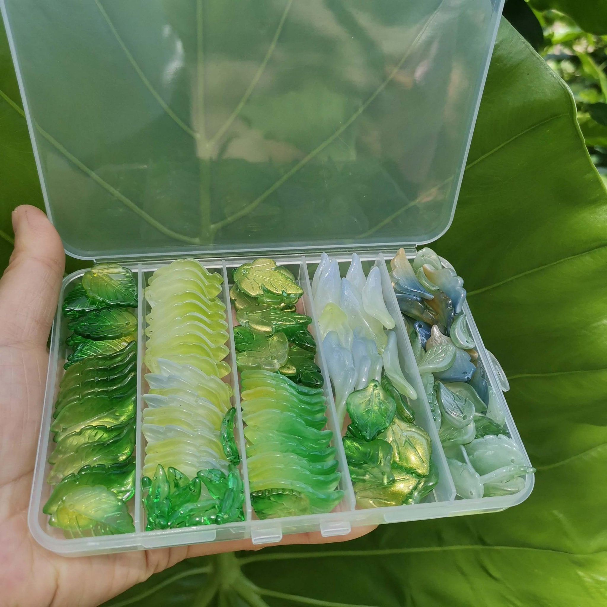 Hand holding a clear plastic organizer filled with assorted green and blue glass leaf beads for jewelry making.