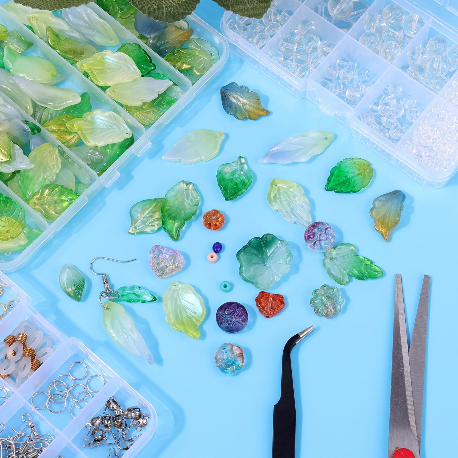 Assortment of Czech glass leaf beads in various green and clear shades, scattered with jewelry making tools on a blue background.